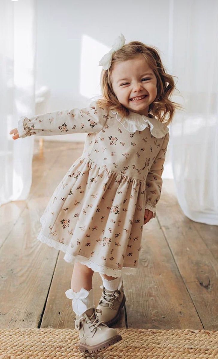 Smiling toddler girl in floral dress with lace collar, bow hair clip, and tan boots dancing on hardwood floor.