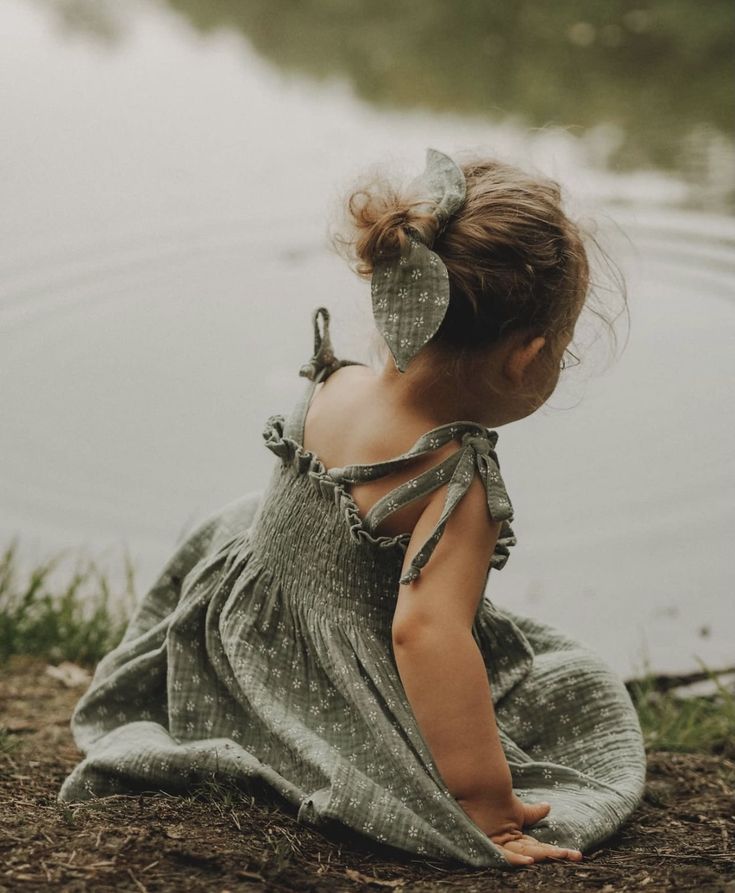 A toddler girl in a sage green smocked dress with a bow in her hair sits by a calm lake.
