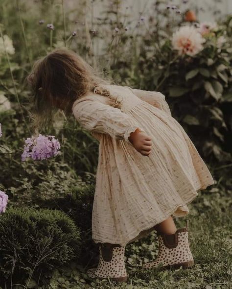 A young girl in a cream floral dress and spotted boots explores a lush garden filled with pink and peach blooms.