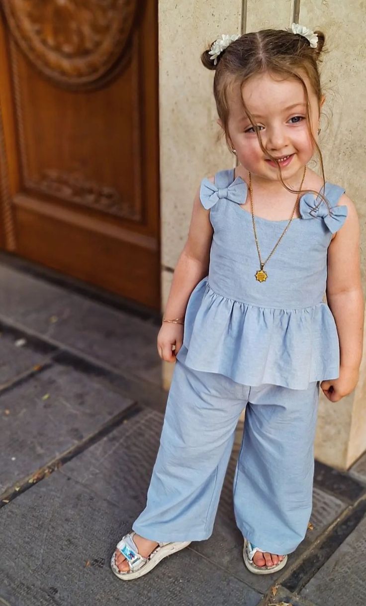 Smiling toddler girl in light blue ruffle top and wide-leg pants with flower necklace standing by wooden door.