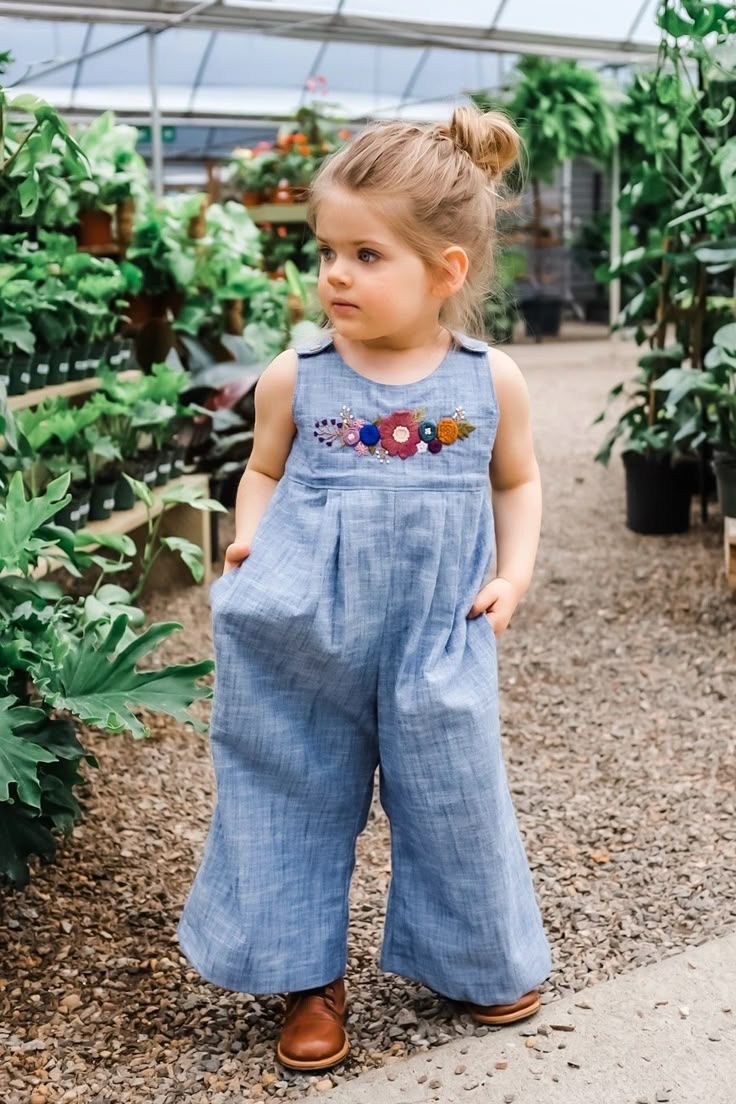 Toddler girl in blue floral embroidered wide-leg jumpsuit posing in a lush greenhouse nursery.