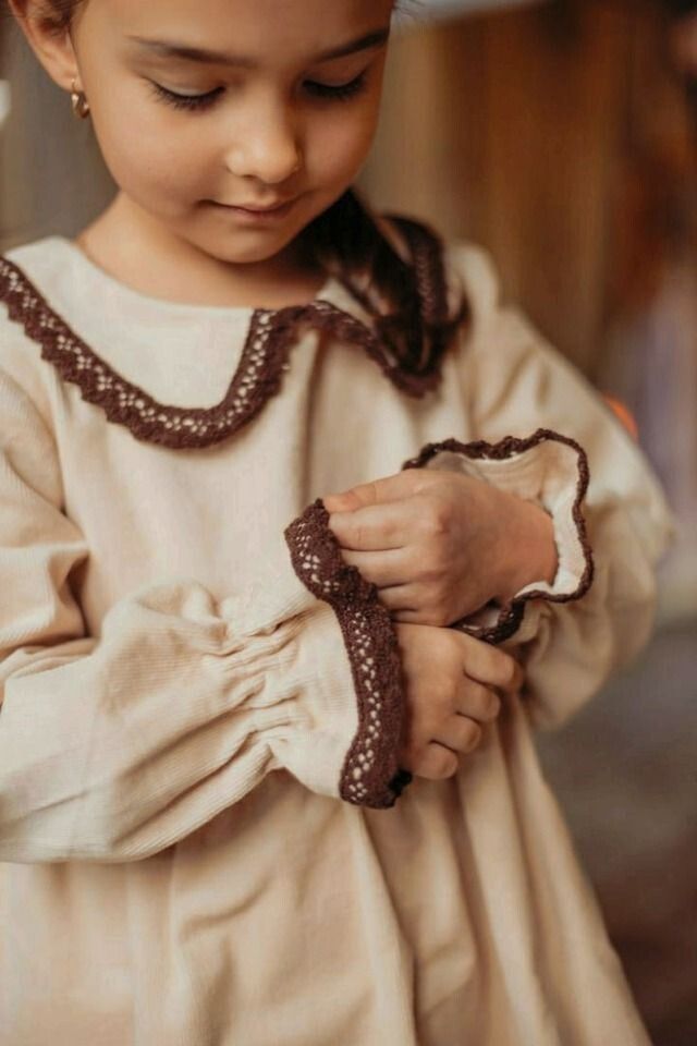 Young girl in beige dress with brown lace trim cuffs and collar, looking down at her sleeve.