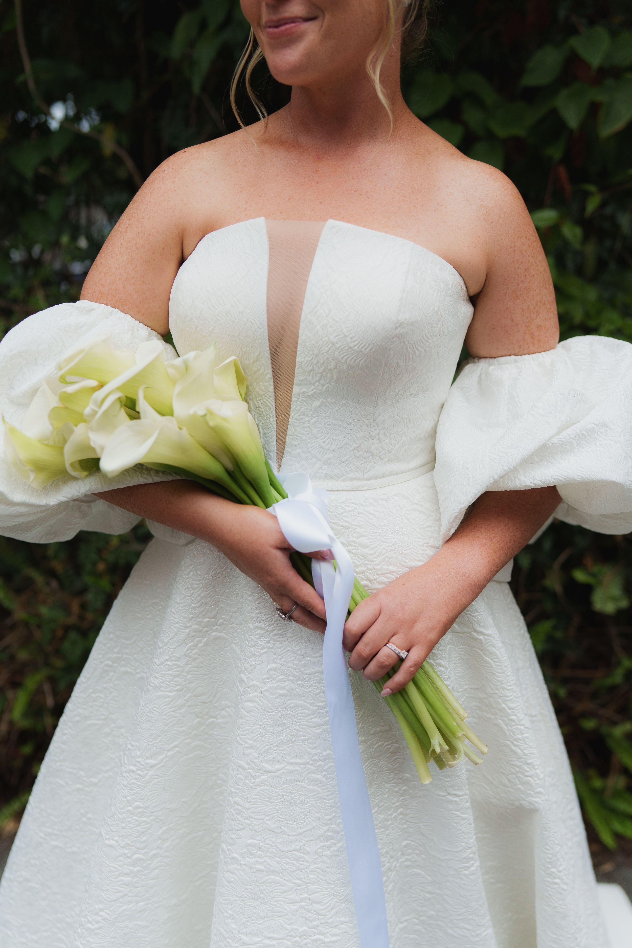 Bride in white strapless gown with puff sleeves holding calla lily bouquet tied with white ribbon outdoors.
