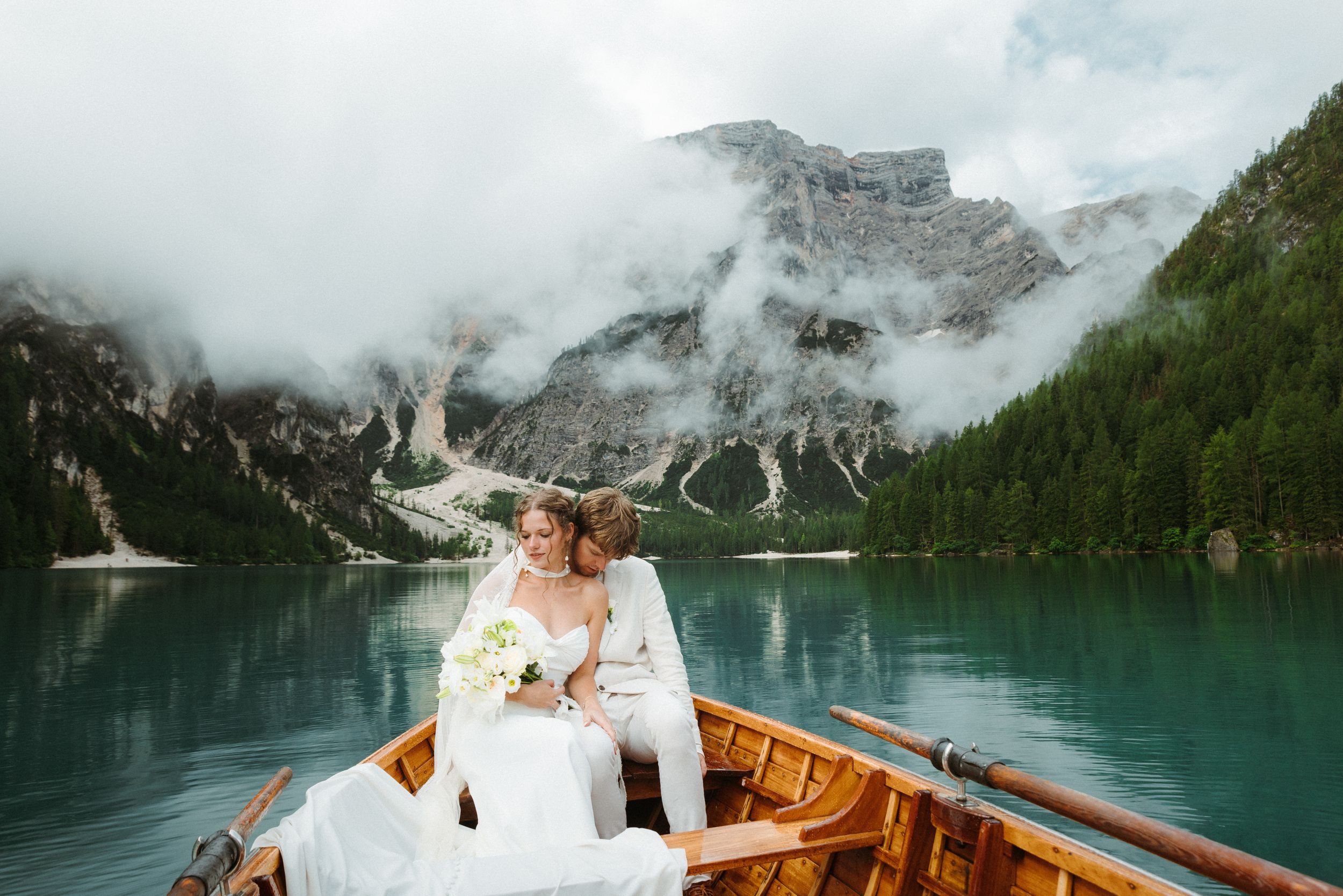 A couple in wedding attire enjoys a romantic boat ride on a turquoise alpine lake surrounded by misty mountain peaks.