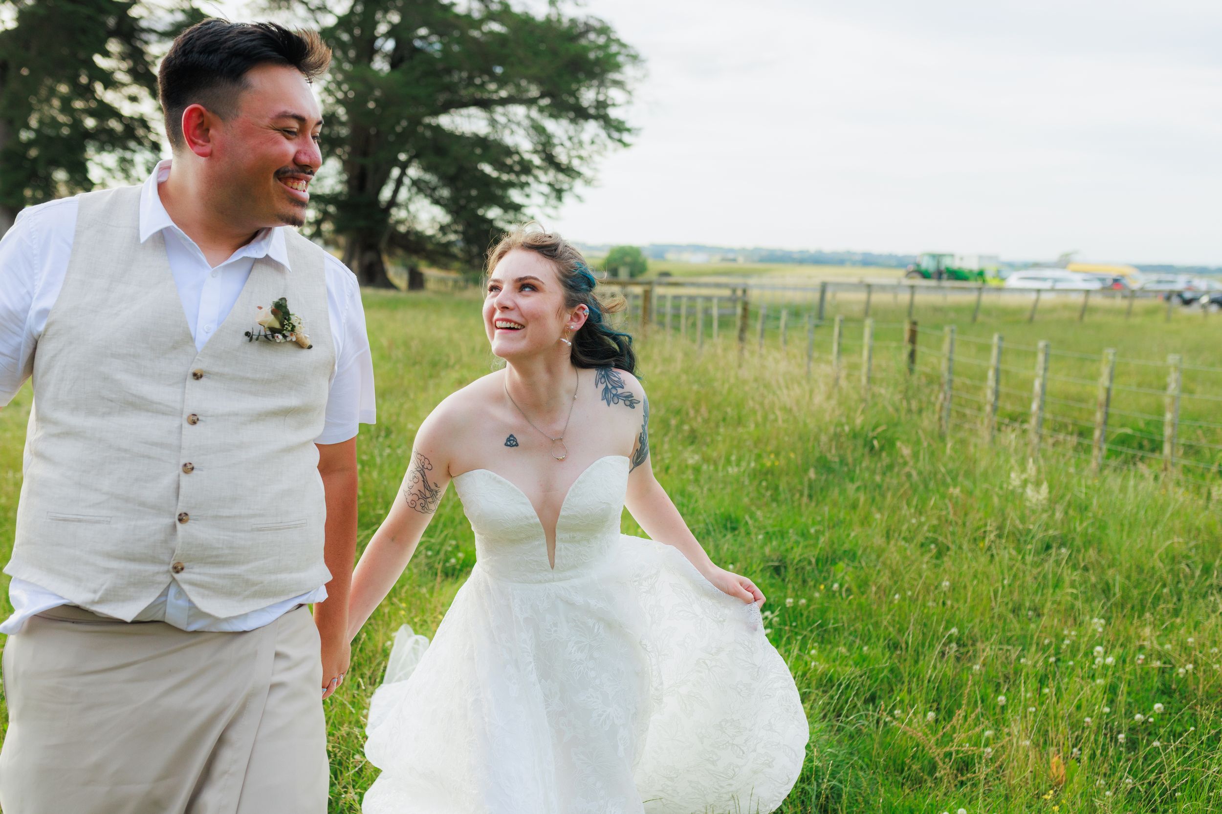 A couple in wedding attire runs playfully through a lush green meadow on a sunny summer day.