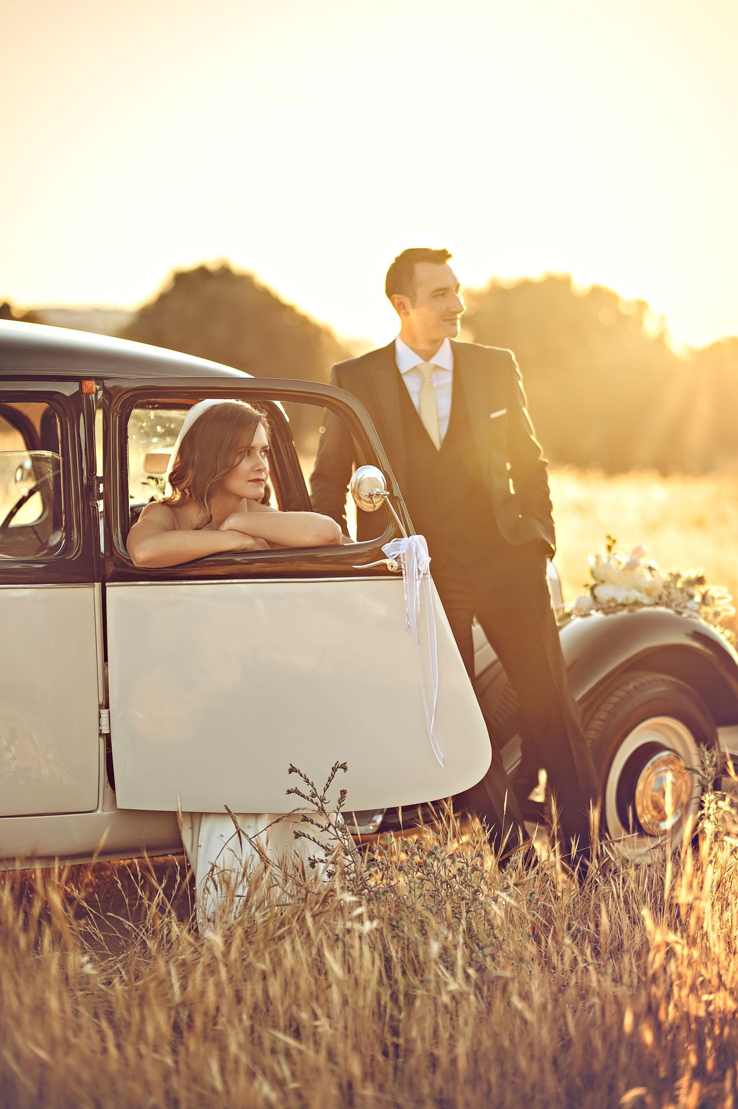 Newlyweds pose with classic car in field during golden hour.