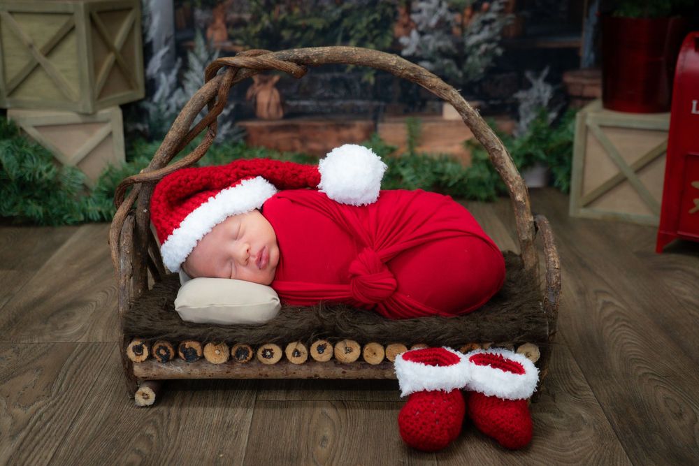 A sleeping baby in red Christmas outfit rests in a rustic wooden basket with matching red booties on the floor.