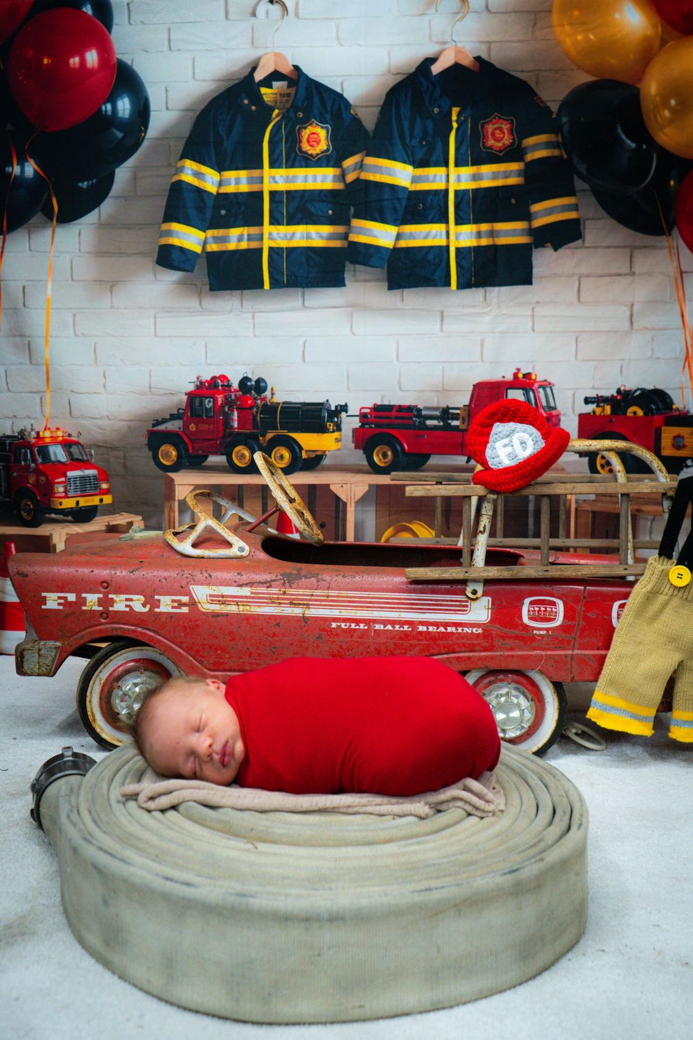 Firefighter themed newborn photo with toy trucks, uniforms on wall, and baby sleeping on coiled fire hose.