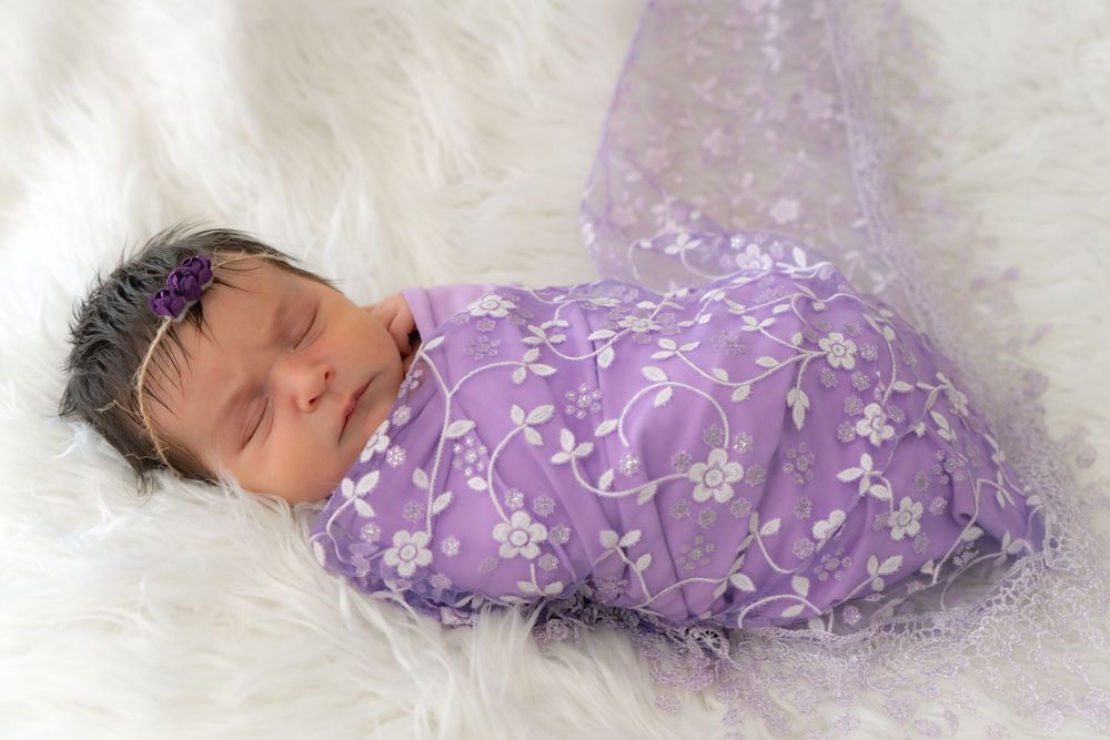 Sleeping newborn wrapped in purple floral swaddle blanket laying on white fluffy background.