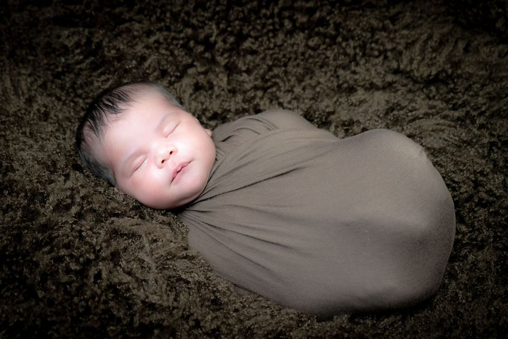 Newborn sleeping peacefully while wrapped snugly in gray swaddle blanket against dark textured background.