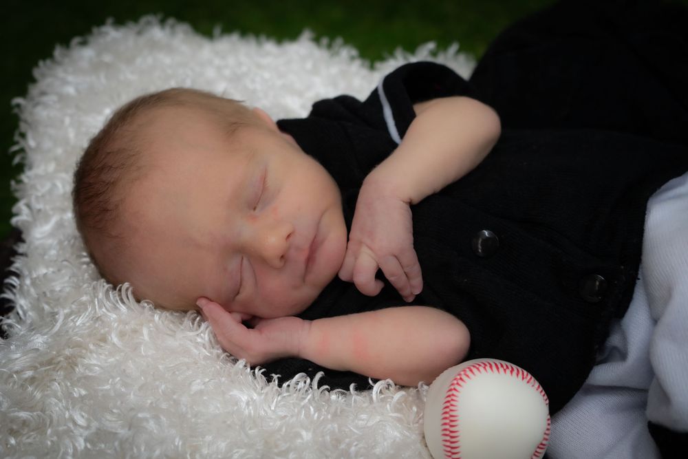 A peaceful sleeping infant in black clothing rests on a white fluffy blanket next to a baseball.