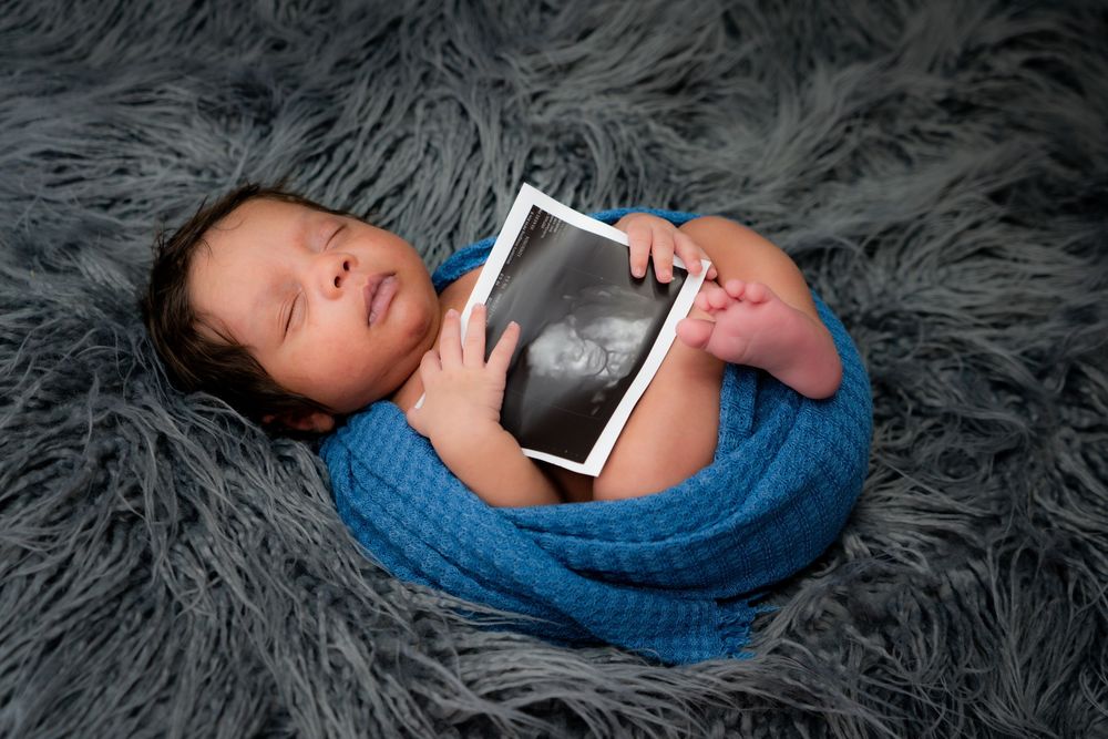 Newborn baby wrapped in blue swaddle holds sonogram photo while lying on gray fur blanket.