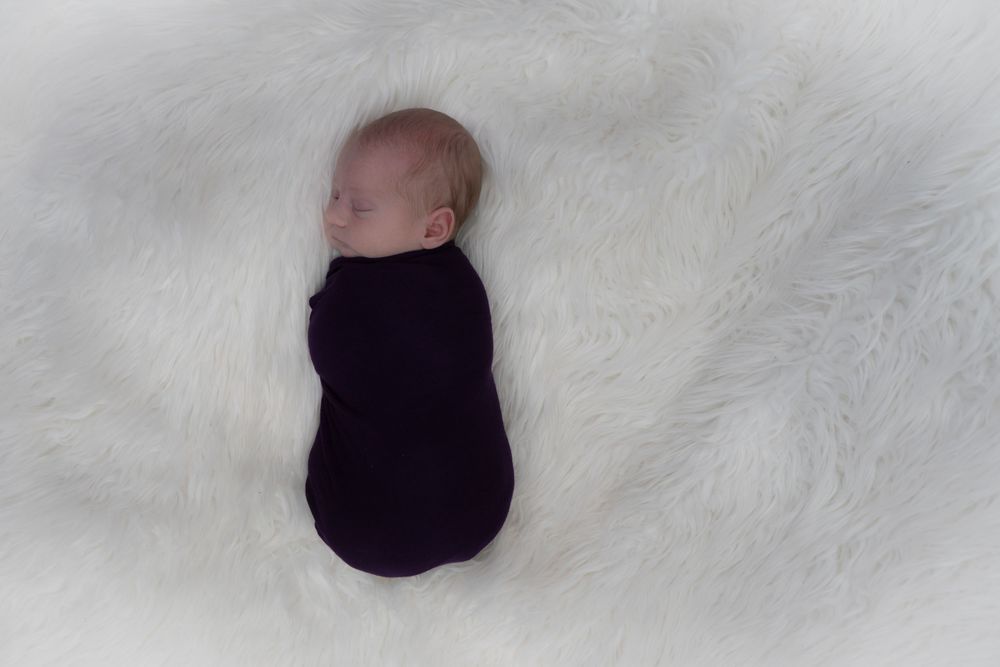A sleeping newborn swaddled in dark purple fabric lies peacefully on a white fluffy blanket.