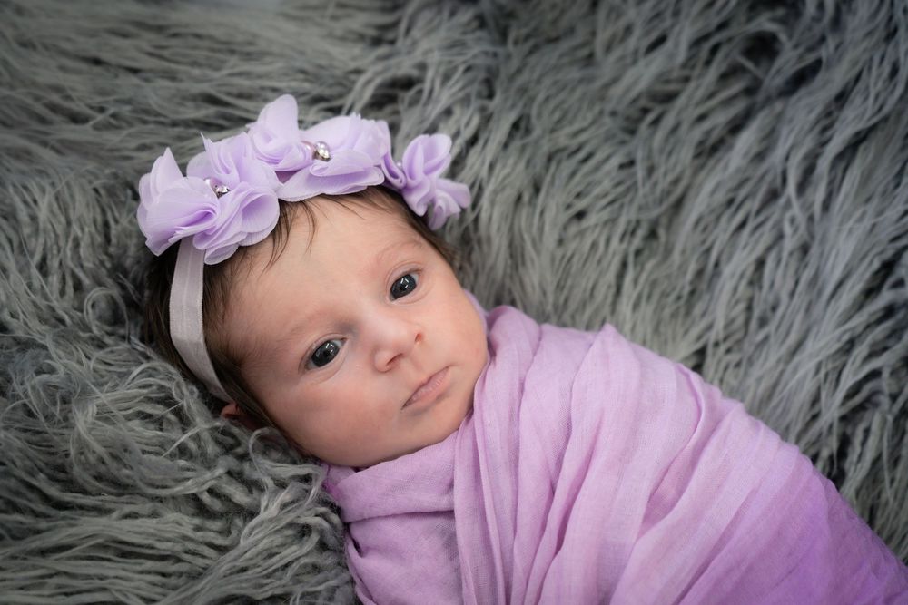 Baby wearing purple floral headband and wrap lies on soft gray fur backdrop during newborn photo session.