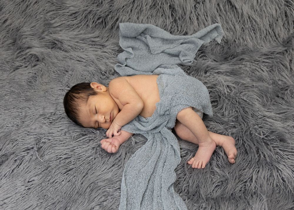 Sleeping baby wrapped in soft gray cloth lying on plush gray fur blanket during newborn photoshoot.