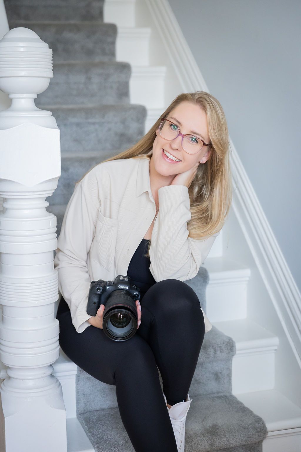 Professional photographer sitting on white stairs holding a DSLR camera while wearing glasses and casual attire.