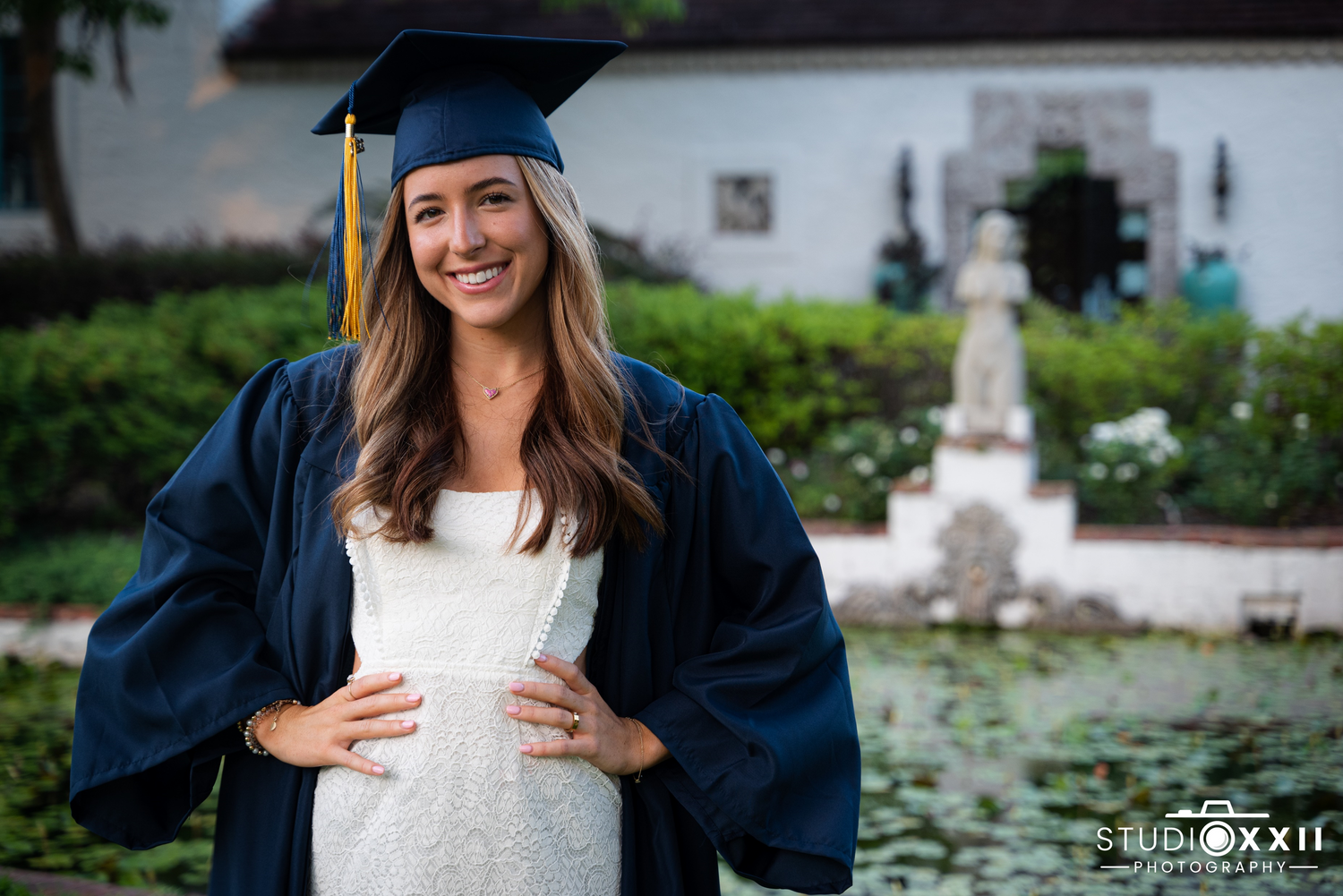 Graduate in blue cap and gown smiling outdoors on graduation day near a garden fountain.