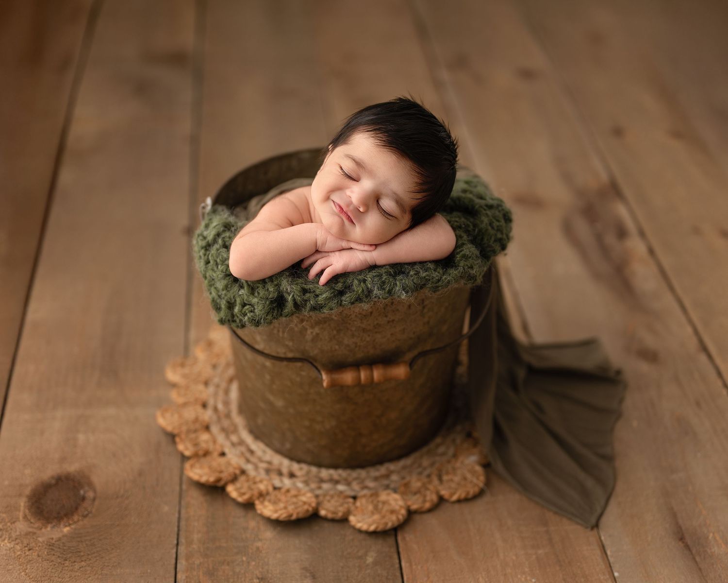 Sleeping newborn baby posed in a rustic wooden bucket with green moss on a warm brown wood floor.