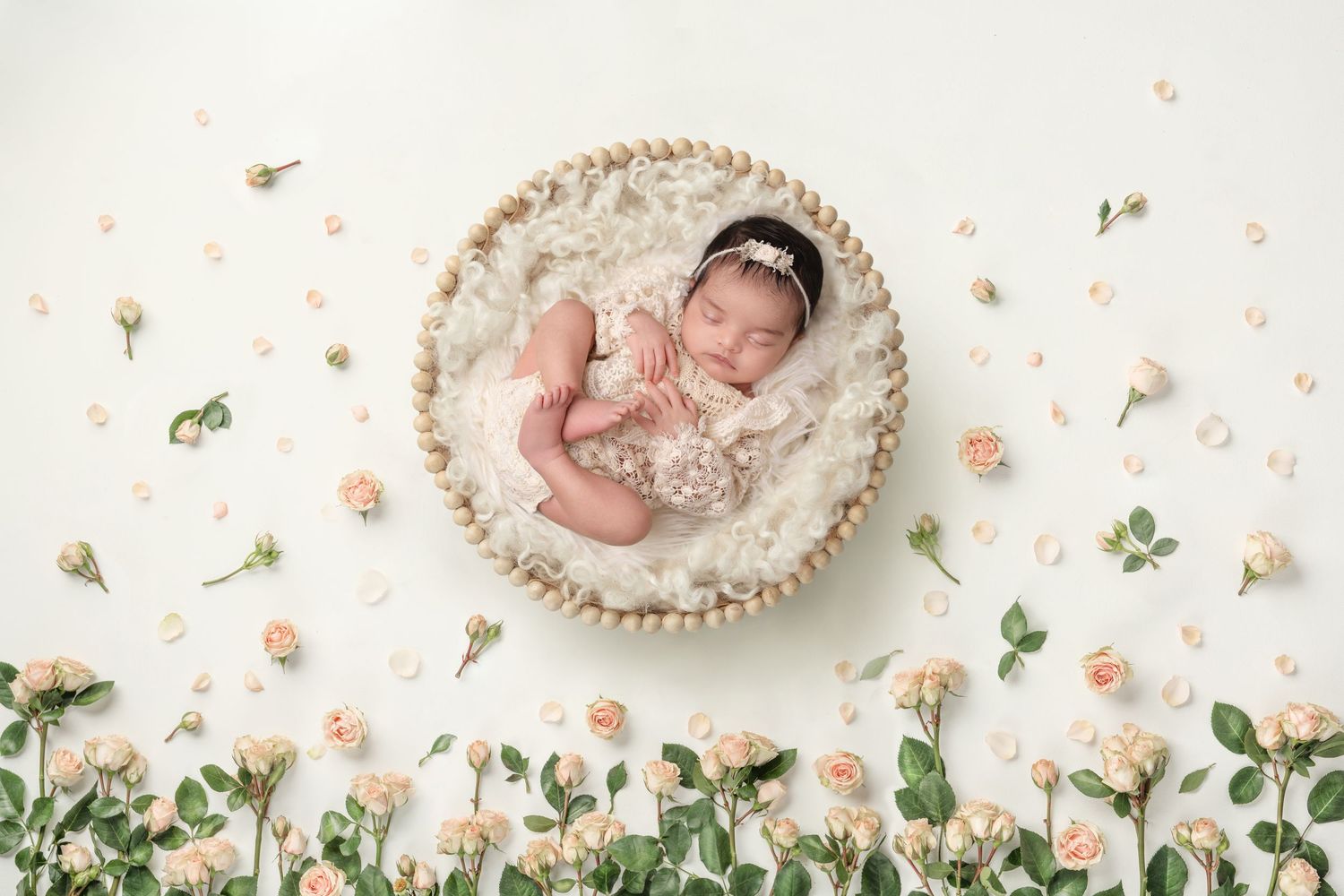 Delicate newborn photo setup with baby in basket surrounded by scattered pink flowers and greenery.