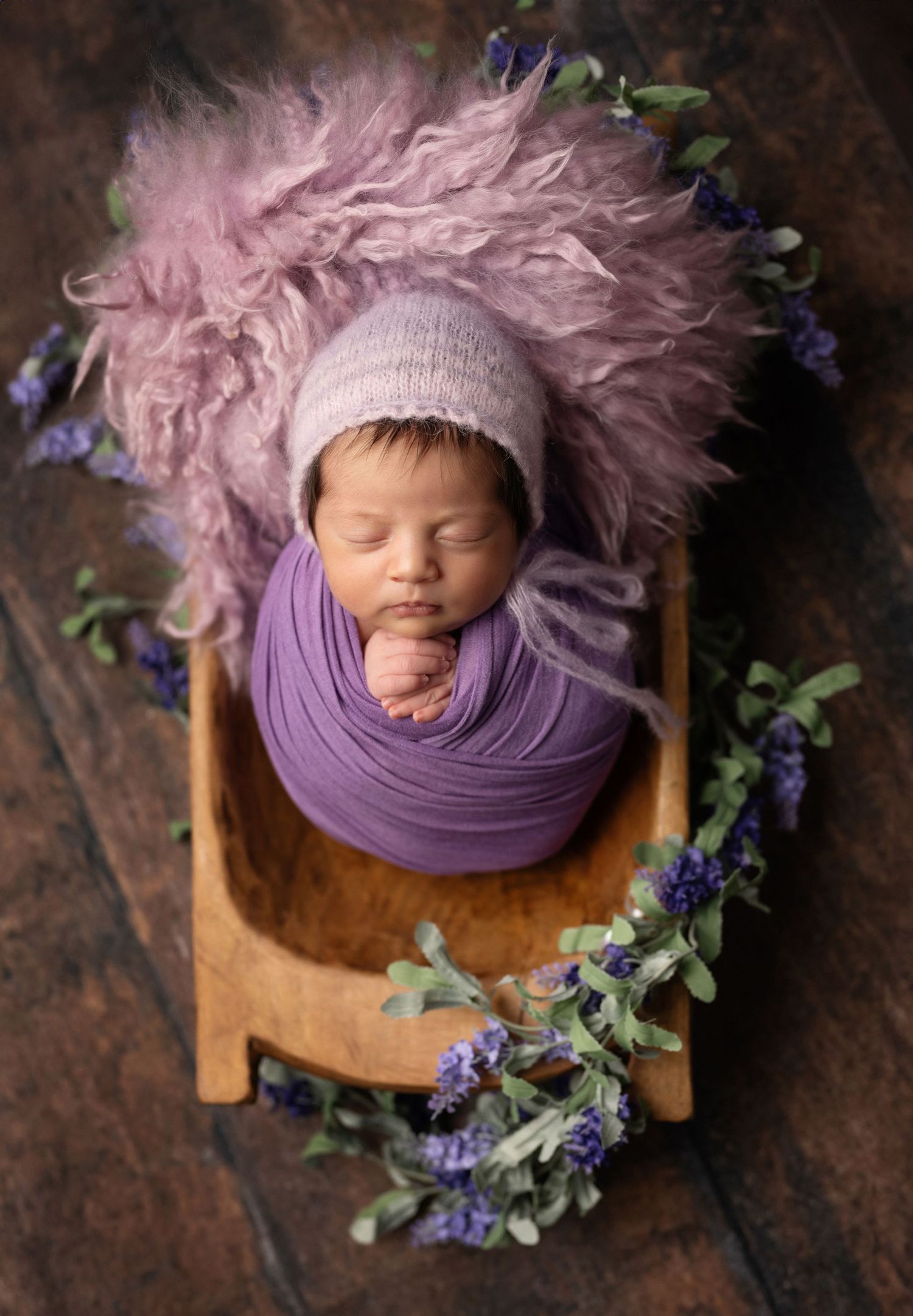Newborn photography featuring a sleeping baby wrapped in purple fabric with delicate floral decorations.