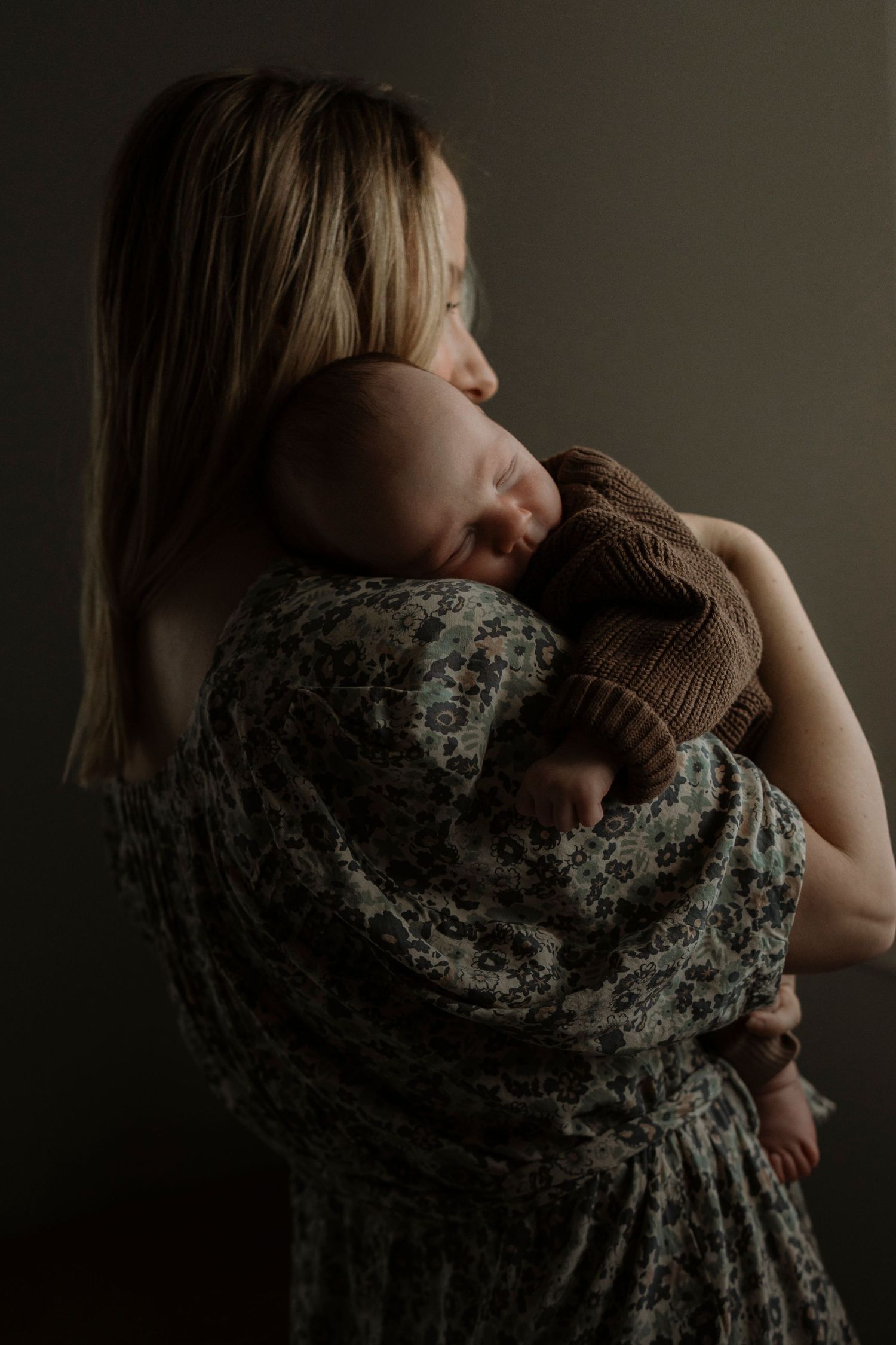 A tender moment captured as a mother in a floral dress cradles and holds her sleeping newborn baby in moody lighting.