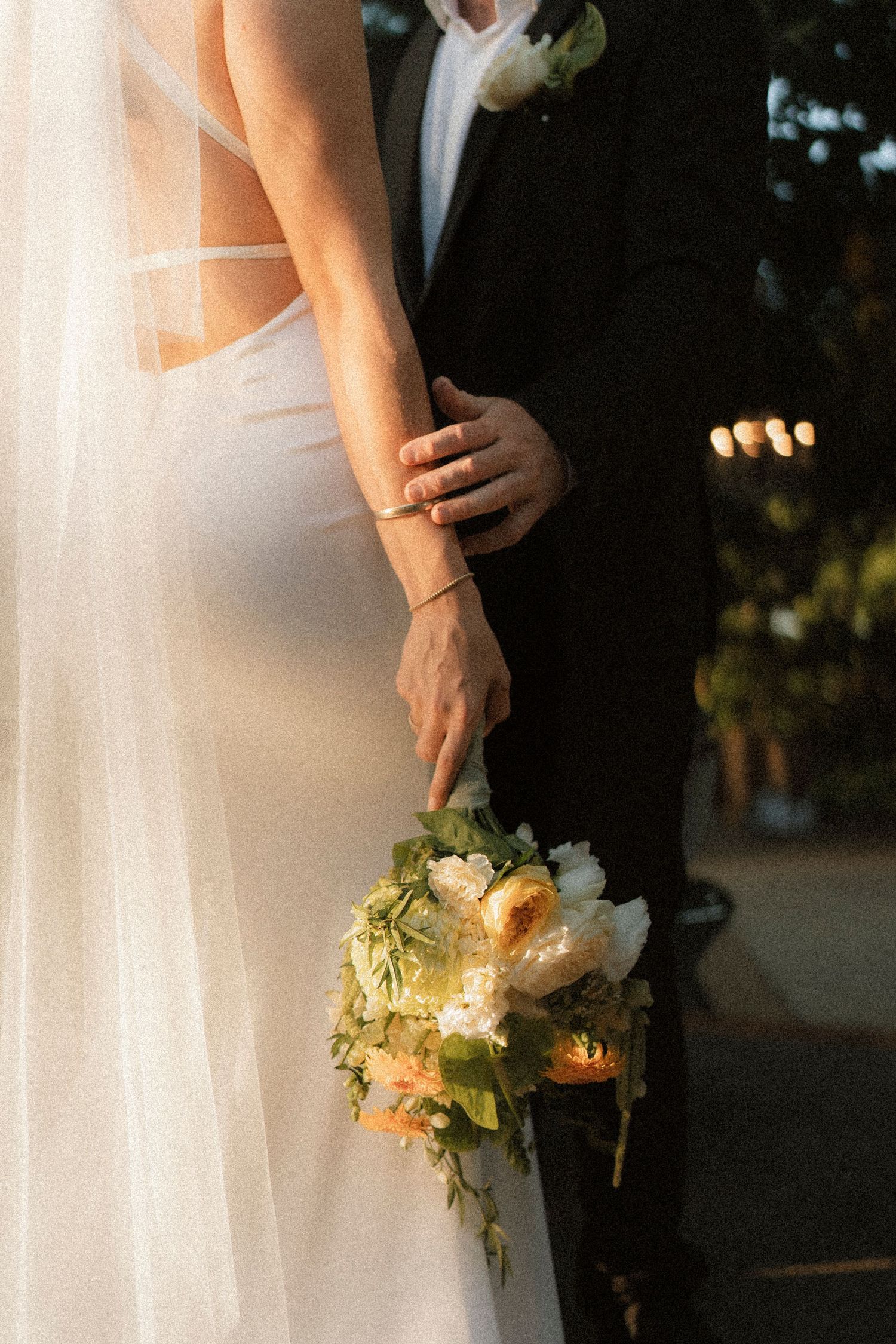 Bride and groom standing close together, bride holding a white and green wedding bouquet in golden hour light.