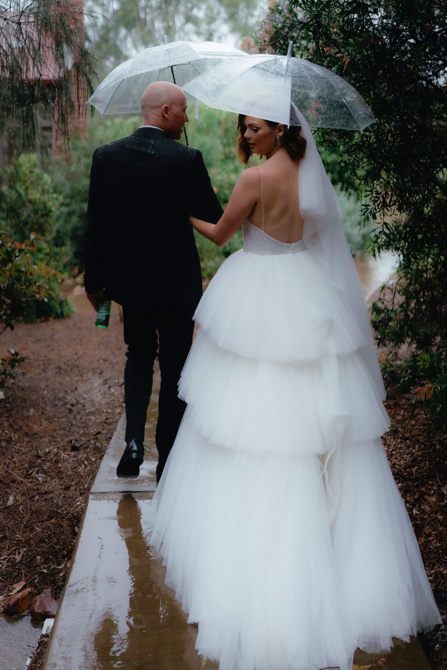 A bride and groom walk together on a wet path through a garden setting during a rainy wedding photo session.
