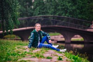 young woman sits in grass field in front of an iron bridge