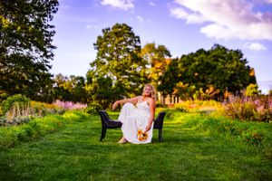 a garden is the scene in this senior portrait of a girl dressed in white sitting on a black velvet couch