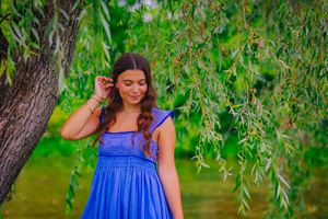 Young woman in blue dress stands underneath a weeping willow tree