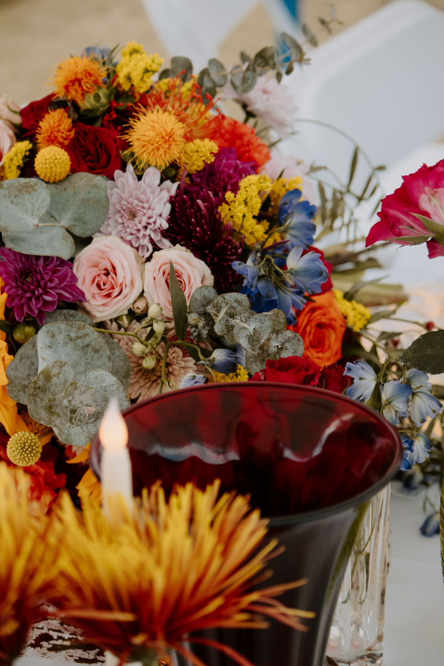 Vibrant wedding floral centerpiece with roses, dahlias, and eucalyptus in red, yellow, pink, and blue beside a dark red vase.