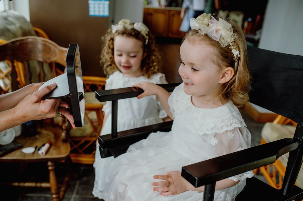 Two young girls in white dresses and floral headbands sit in chairs, one holding a mirror as they prepare for a photoshoot.