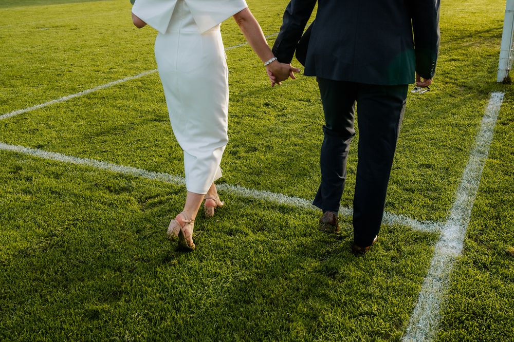 A newlywed couple walks hand in hand on lush green grass, their shoes slightly muddy, following a white chalk line on the field. Roscommon.