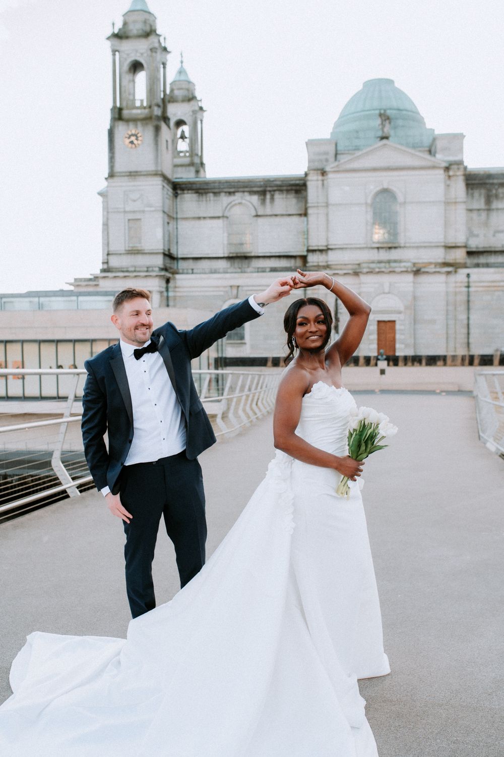 Elegant bride in strapless white wedding gown holding bouquet twirls with groom in black tuxedo on urban bridge, Athlone, Ireland.