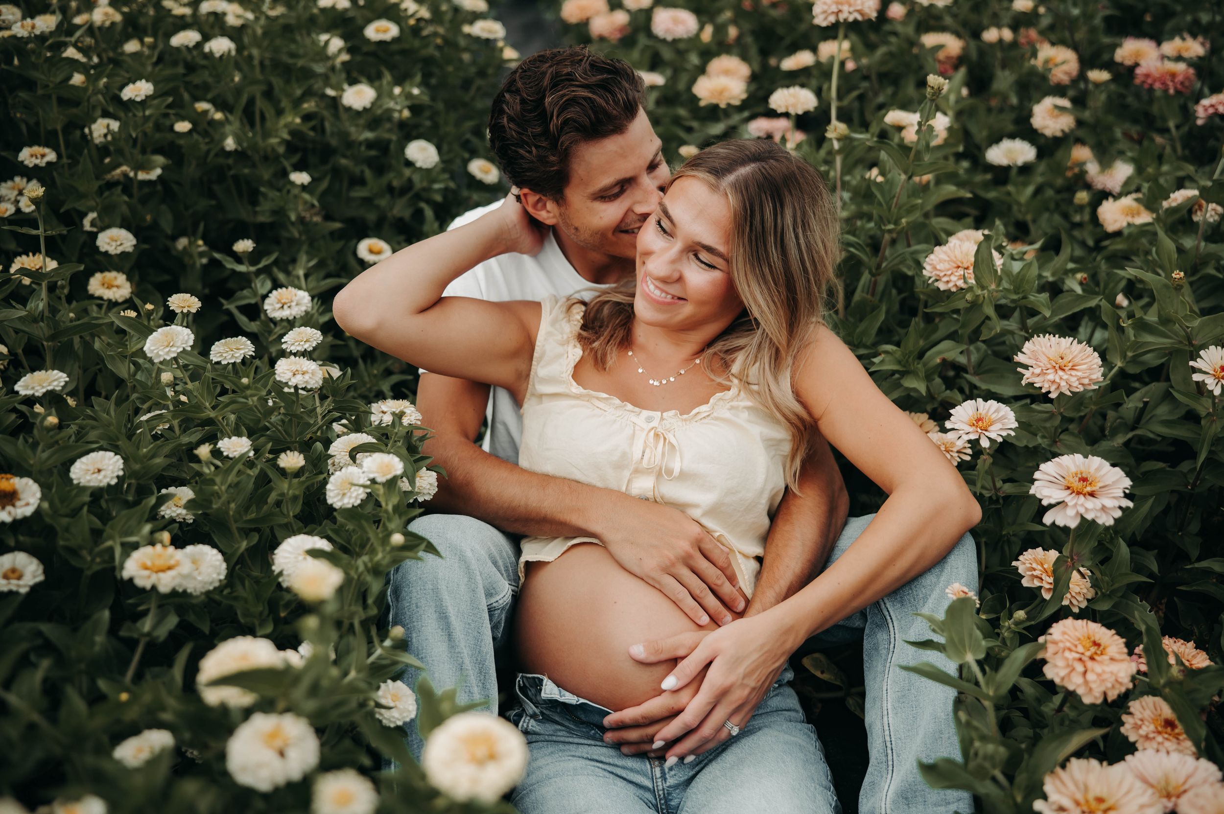 Couple sitting in a flower field, man kissing pregnant woman's head while holding her baby bump.