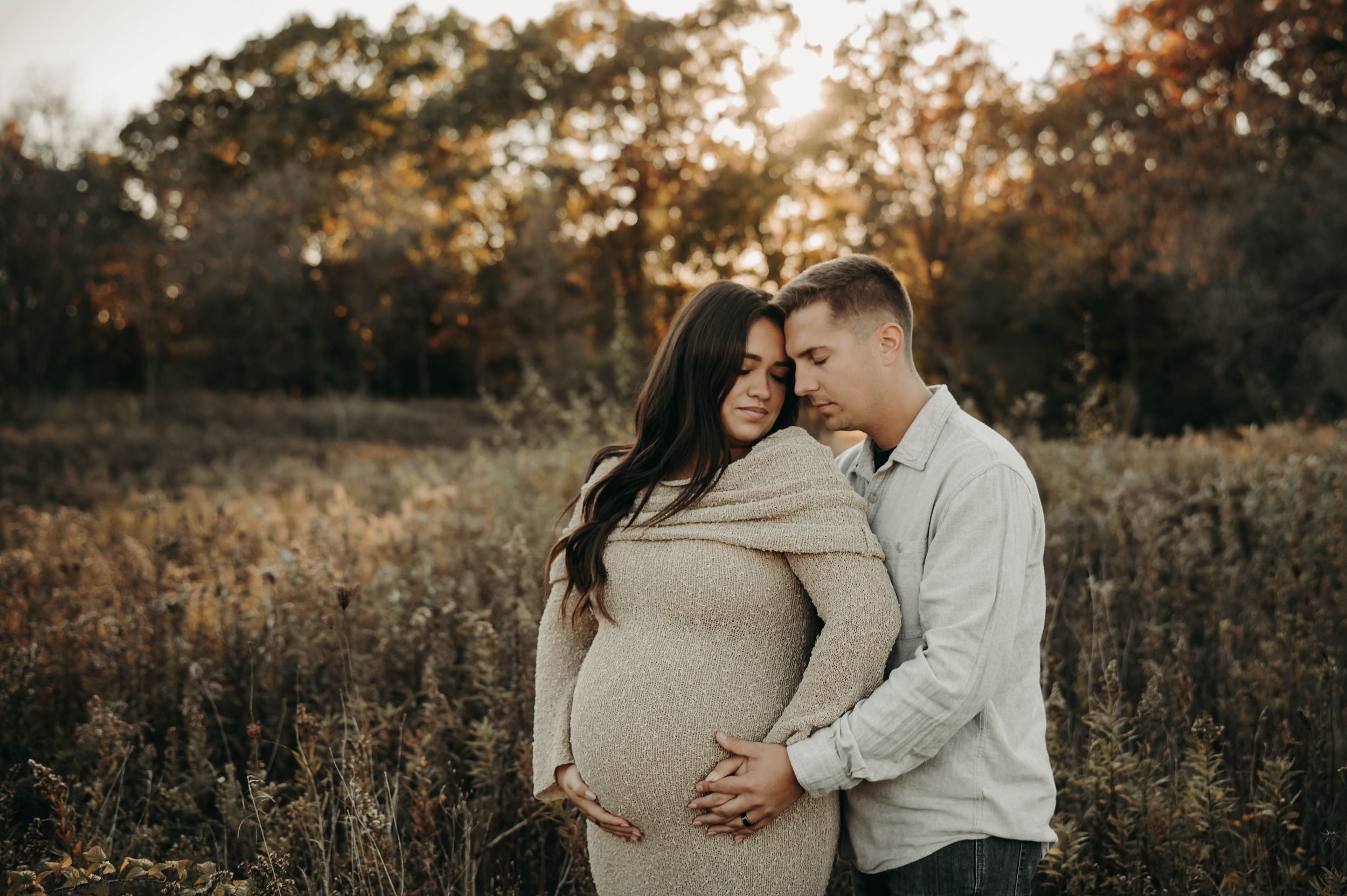 Couple embracing in golden autumn field during maternity photo session at sunset.