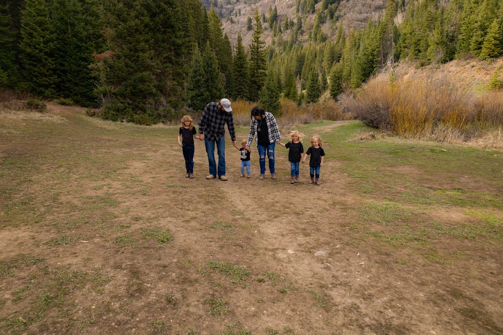 Family of six walking hand-in-hand through a scenic mountain meadow surrounded by evergreen trees in autumn.