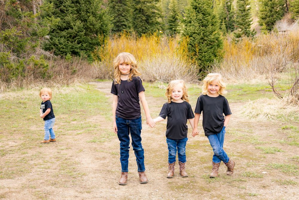 Four blonde children in matching black shirts and jeans hold hands while walking on a dirt path surrounded by trees.