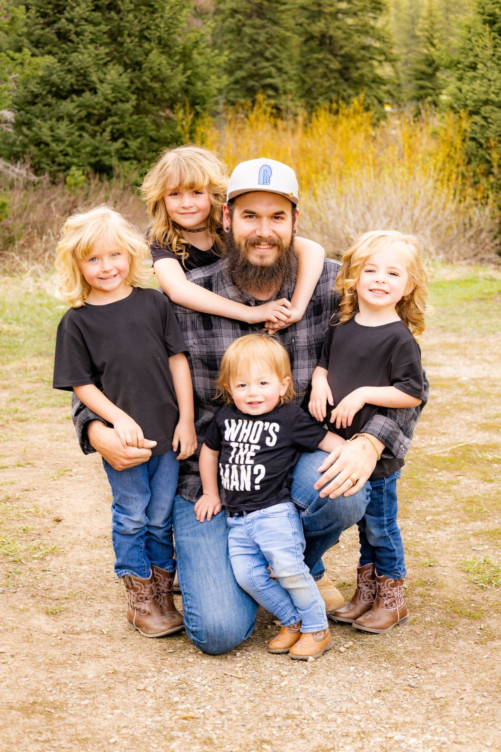 Bearded dad kneeling outdoors with four young blonde children, surrounded by autumn foliage in a mountain setting.