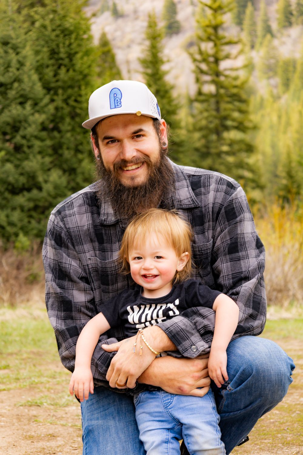 Bearded man in plaid shirt and snapback hat smiling with a toddler on his lap outdoors near pine trees.