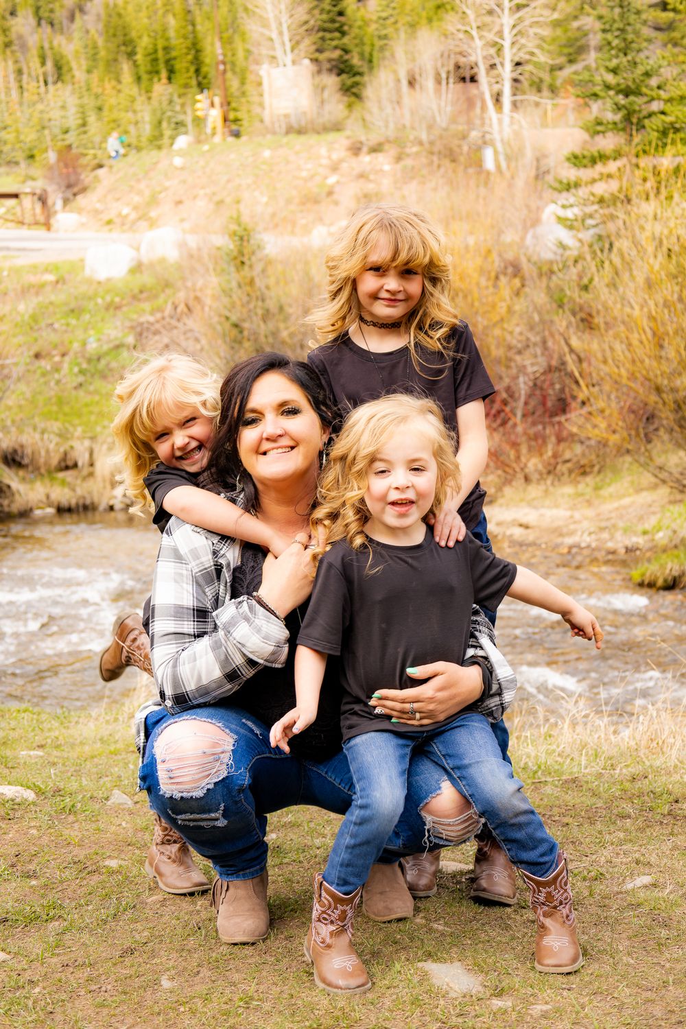 A smiling mother crouches outdoors with her three young blonde children near a mountain stream in Colorado.