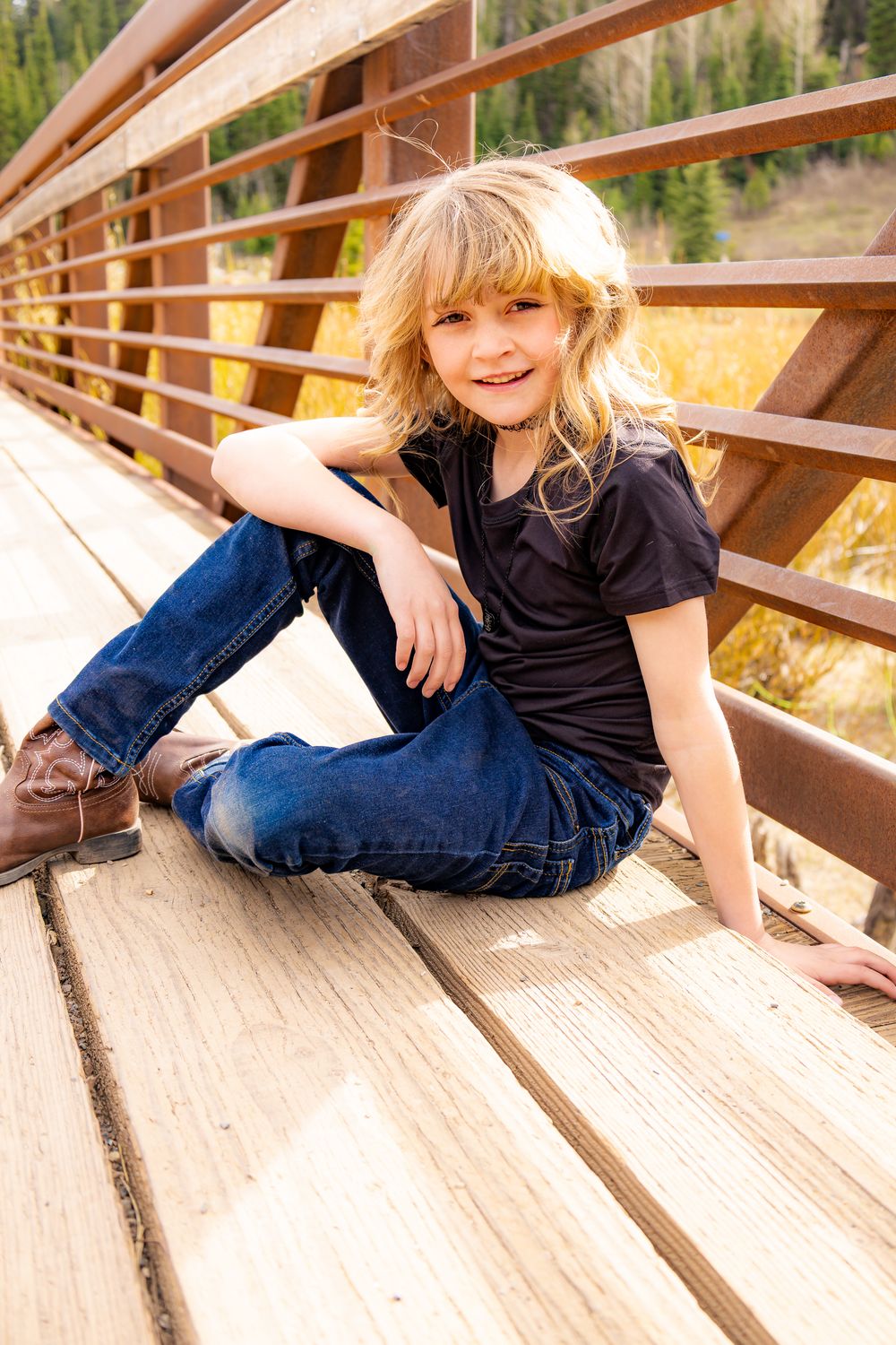 Smiling blonde girl sitting cross-legged on a wooden bridge wearing jeans and black shirt with mountains behind.