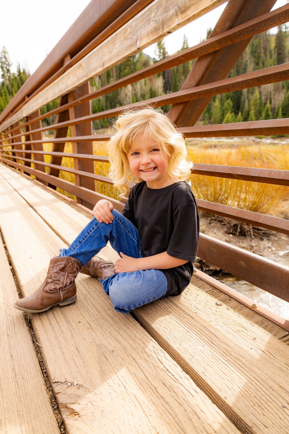 Smiling blonde child sitting on a wooden bridge wearing jeans, black tee, and brown cowboy boots with fall foliage behind.