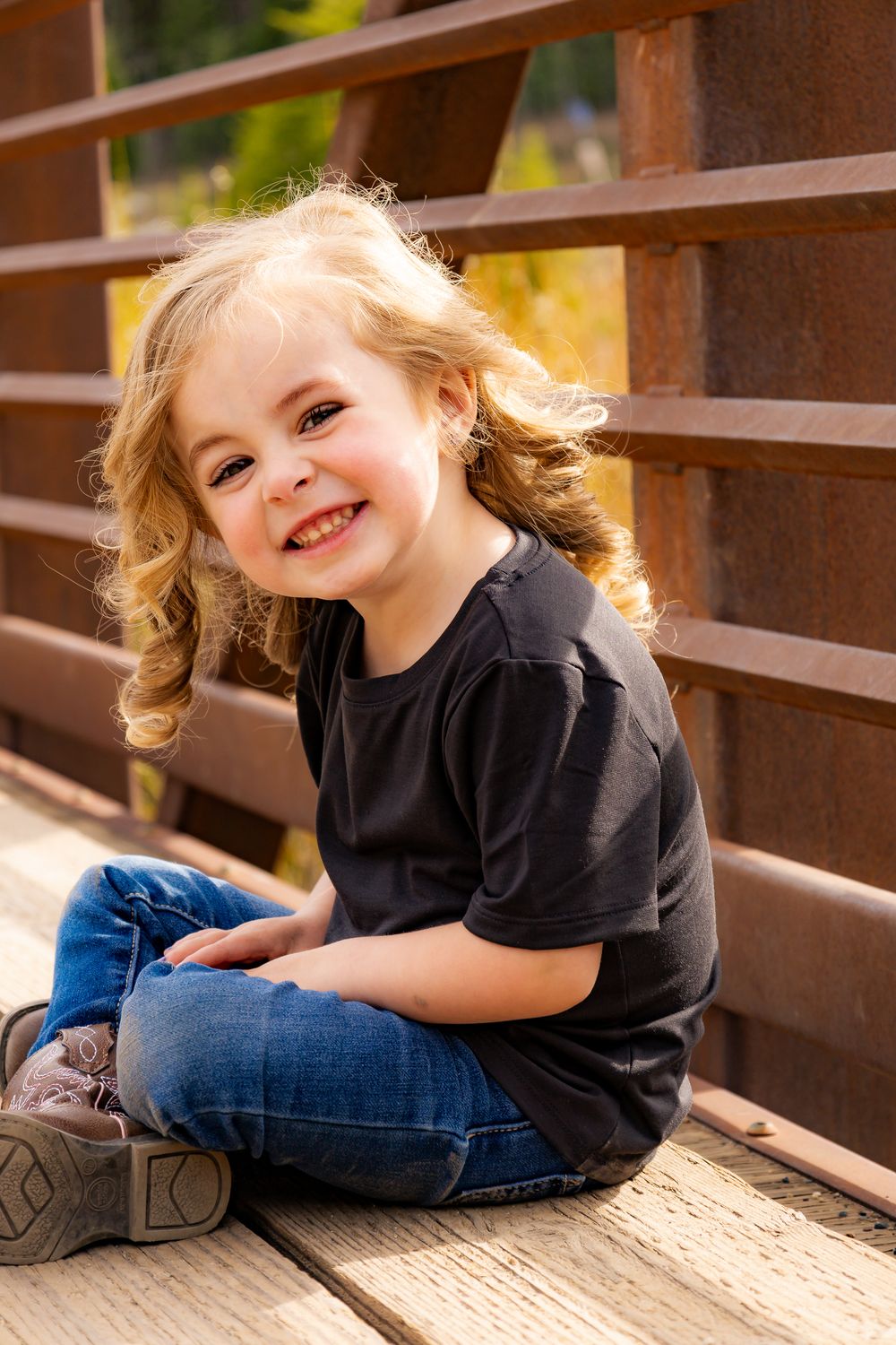 Smiling young girl with curly blonde hair sitting cross-legged on a wooden bridge wearing a black t-shirt and jeans.