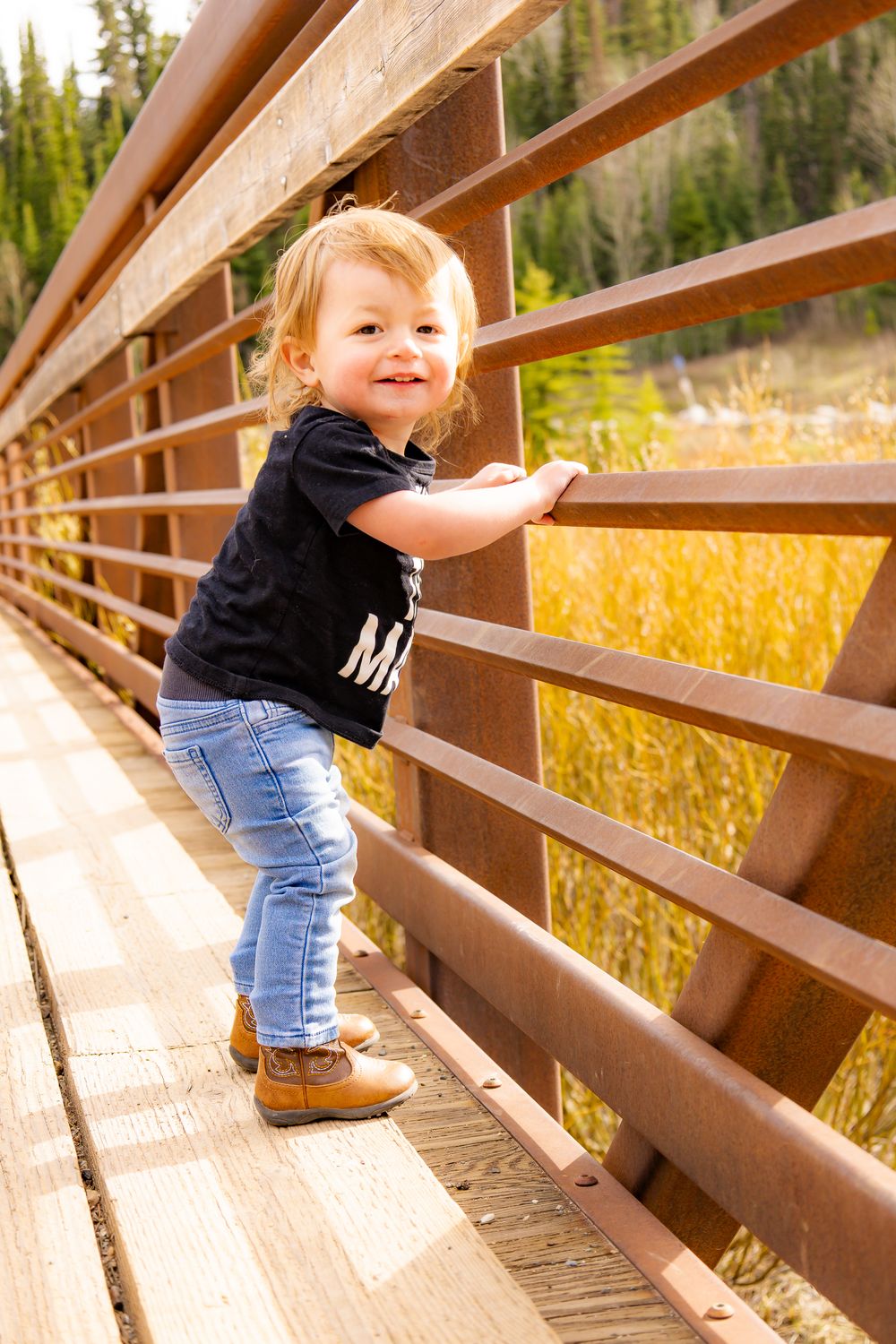 Toddler in black shirt and jeans smiling on a rustic wooden bridge surrounded by autumn foliage.