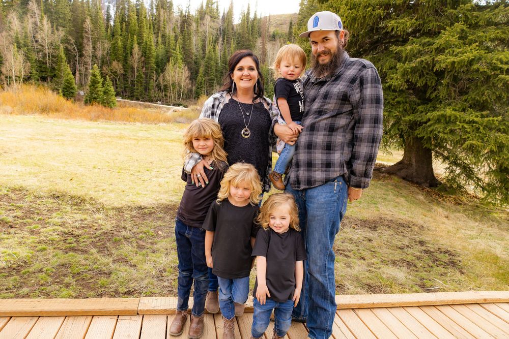 Happy family of six posing on a wooden deck surrounded by pine trees and golden autumn meadows in a mountain setting.