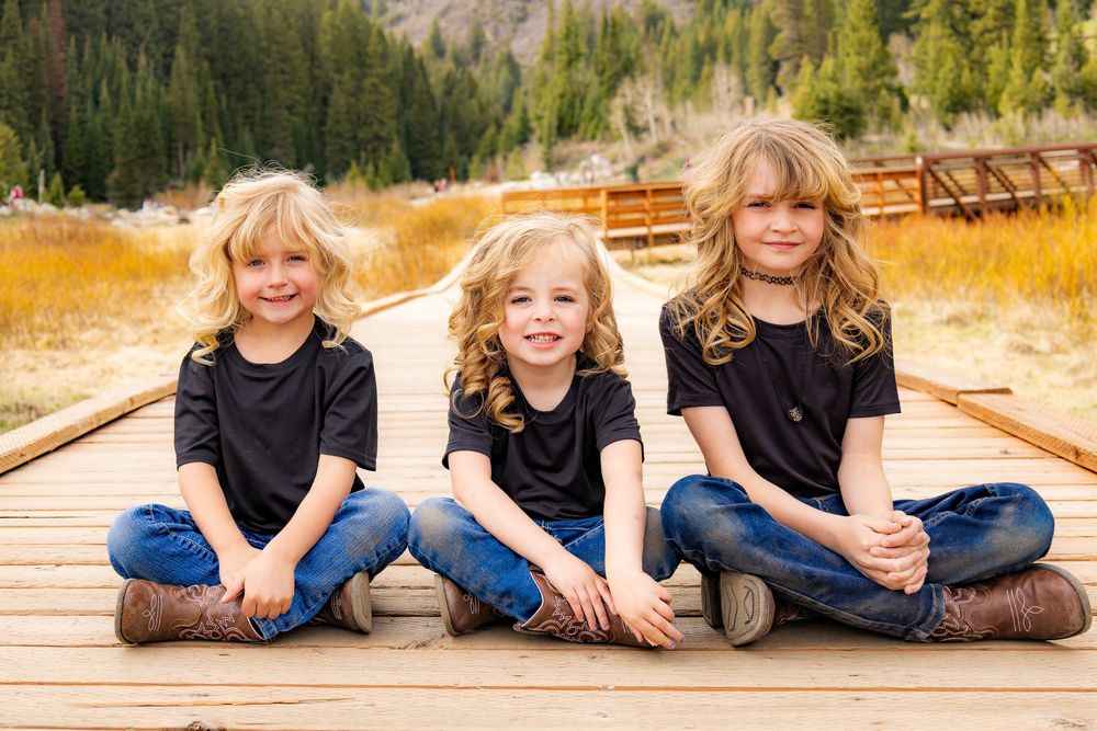 Three blonde siblings in matching black shirts and jeans sitting cross-legged on a wooden path outdoors.