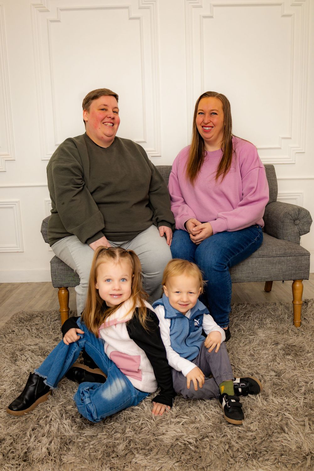 Two women sit on a gray sofa smiling with two young children sitting on a rug in front of them.