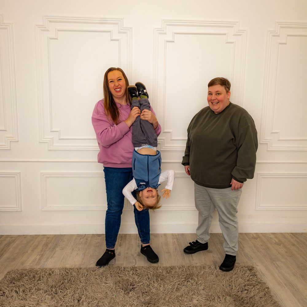 Two women and a child playing indoors, one woman holding a laughing toddler upside down in a bright white room.