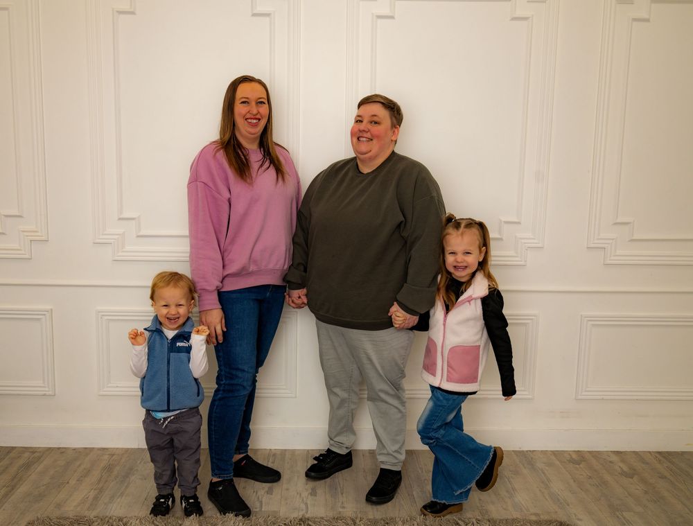 Family of four posing against white decorative wall, two adults with young boy and girl in casual colorful outfits.